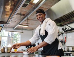Chef traiteur professionnel souriant préparant un repas de mariage dans une cuisine moderne des Caraïbes, portant une toque et un tablier.
