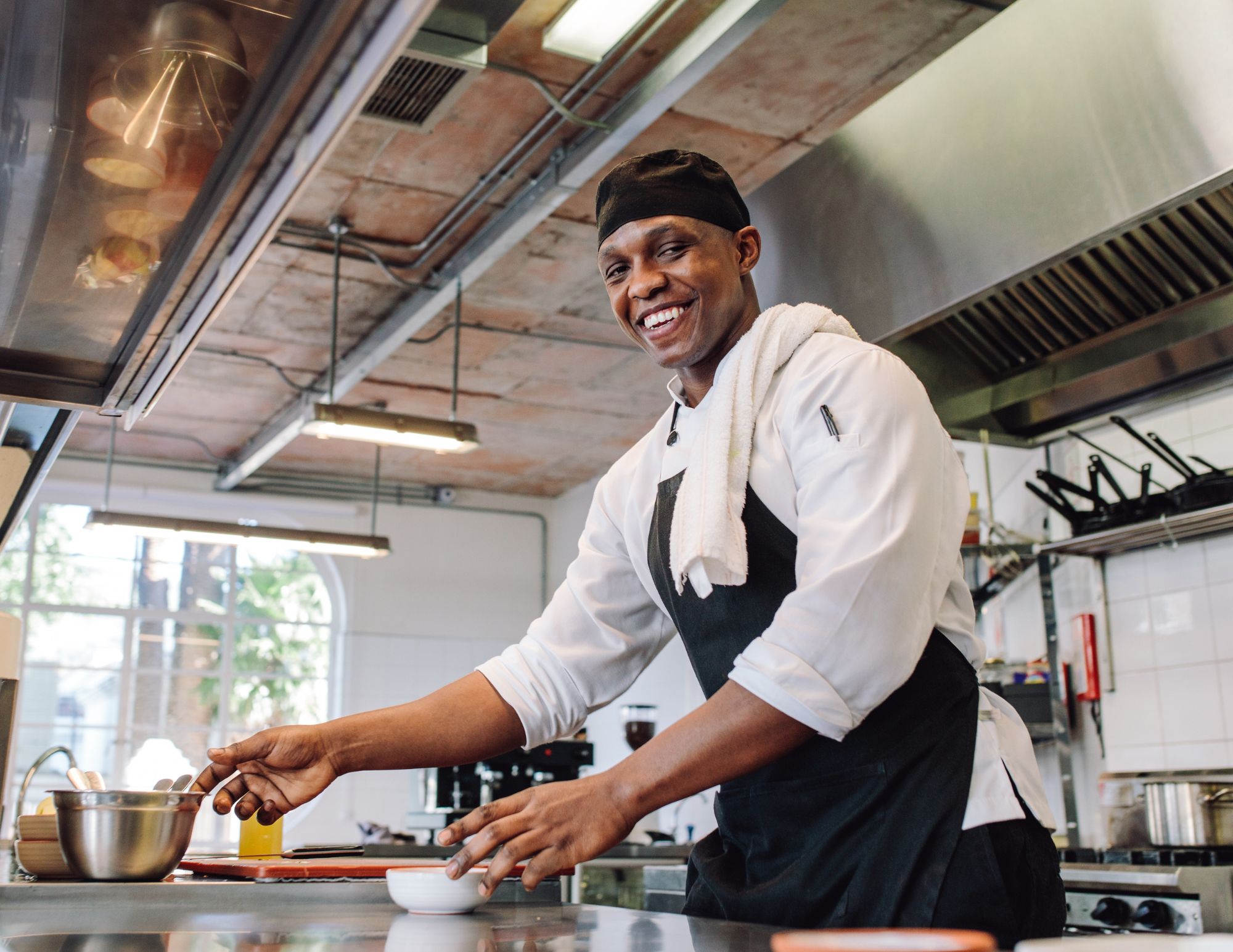 Professional wedding caterer chef smiling while preparing Caribbean wedding meal in modern kitchen wearing black toque and apron.