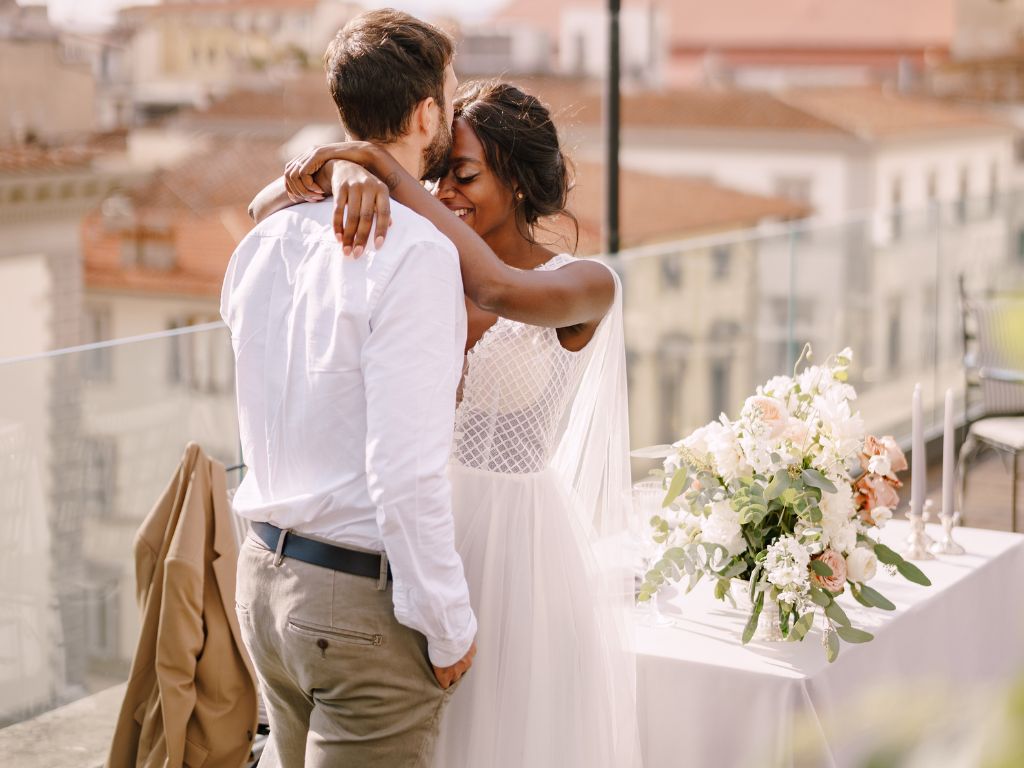 Couple de jeunes mariés s'enlaçant sur un balcon avec vue panoramique, mariage intime en bord de mer aux Caraïbes avec bouquet de fleurs blanches et verdure